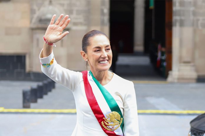 Mexico's President Claudia Sheinbaum smiling and waving at crowds as she stands in front of the National Palace in Mexico City. She is wearing a white dress with traditional embroidery on the cuffs and bearing Mexico's red, white and green presidential sash.