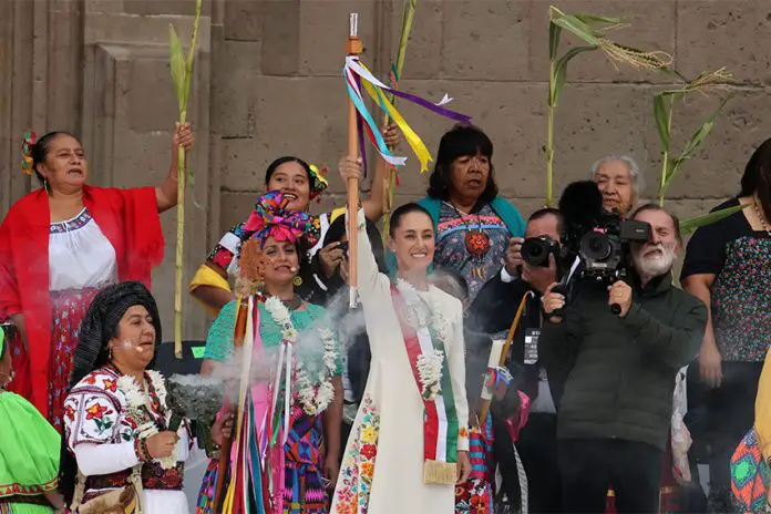 Mexican President Claudia Sheinbaum holding up the ceremonial presidential baton, surrounded by Mexican women, many of them dressed in traditional indigenous clothing. Professional photographers stand nearby taking photos of the event.