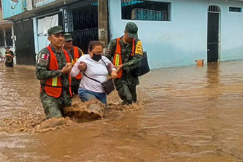 Two soldiers in military fatigues and organe emergency vests hold the arms of a woman in jeans and a white shirt who is carrying a purse as they help her navigate knee-deep muddy floodwaters in Acapulco.