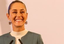 Claudia Sheinbaum staring into the camera at a press conference and smiling.