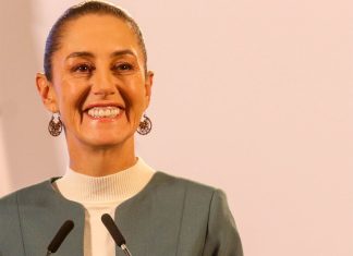 Claudia Sheinbaum staring into the camera at a press conference and smiling.