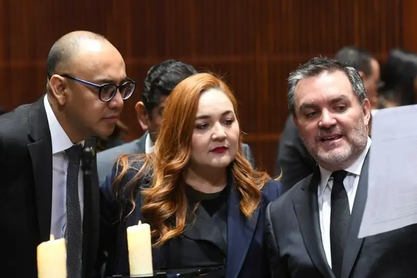 three members of Mexico's Chamber of Deputies in session, standing while one (male) member shows the other two members (a woman and a man) a computer printout.