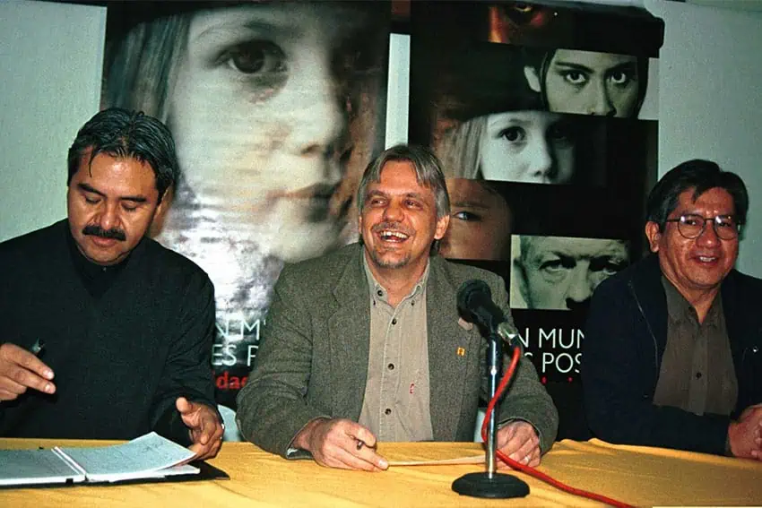 Three men sitting at a table with a microphone at what looks like a press conference. The man in the center, Carlos Imaz Gispert, is seated in front of the microphone and smiling.