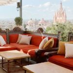 Luxurious sofas made of carved dark wood and with orange cushions and white and beige pillows on a rooftop terrace looking at skyline of San Miguel de Allende, Guanajuato, Mexico.
