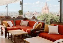 Luxurious sofas made of carved dark wood and with orange cushions and white and beige pillows on a rooftop terrace looking at skyline of San Miguel de Allende, Guanajuato, Mexico.