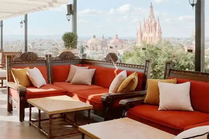 Luxurious sofas made of carved dark wood and with orange cushions and white and beige pillows on a rooftop terrace looking at skyline of San Miguel de Allende, Guanajuato, Mexico.