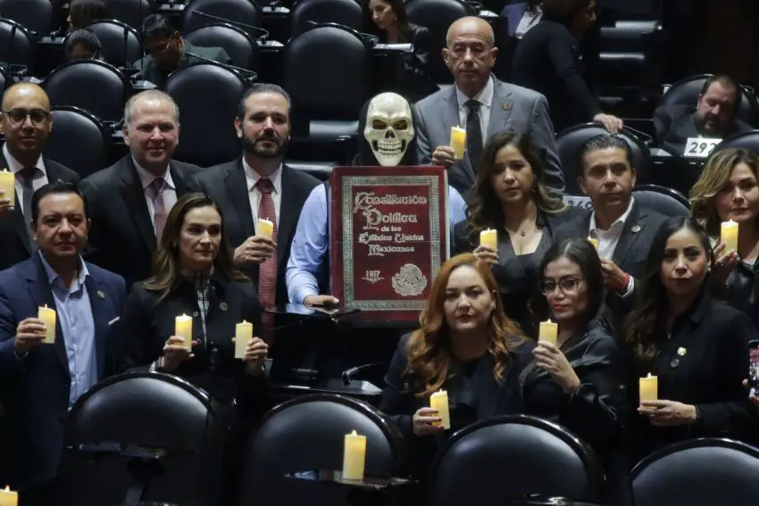 Members of Mexico's Chamber of Deputies holding lit funeral style candles in the Chamber, posing for a picture with a deputy in a dress shirt and vest and a skeleton mask, holding up an ironic mockup of the book of the Mexican constitution