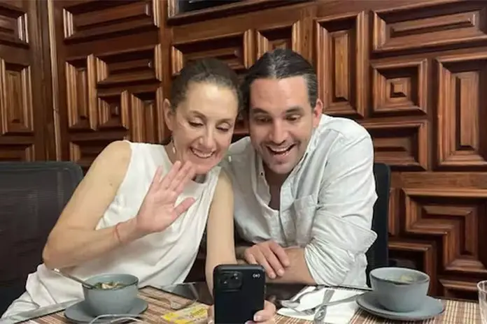 Claudia Sheinbaum and her stepson Rodrigo Ímaz Alarcón, sitting in a restaurant at a table with coffee cups and looking at something on a cell phone