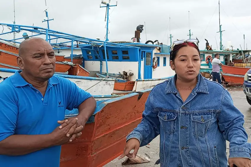 Hombre y mujer de pie en un muelle junto a pequeños barcos pesqueros, mirando a la cámara.