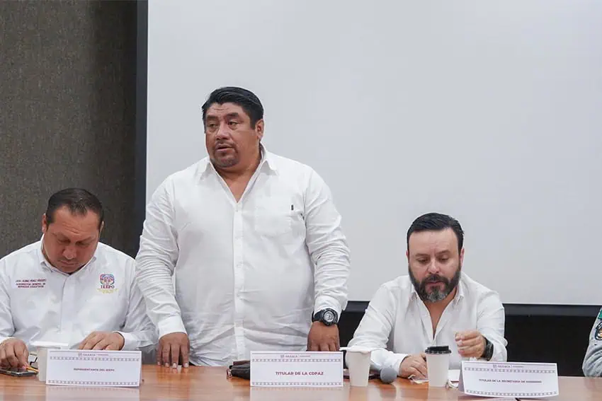 Three Oaxaca state officials sitting at a conference table. At the center, standing, is Donato Vargas.