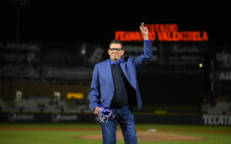 Fernando Valenzuela in a blue suit jacket and jeans holding two fingers up and holding a baseball glove in another hand as he stands inside the Hermosillo Naranjeros baseball team stadium, Estadio Fernando Valenzuela, which is in large red letters in the background of the photo