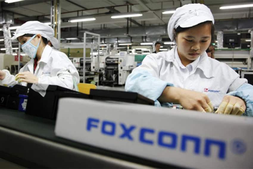 Trabajadores chinos en una fábrica de productos electrónicos de Foxconn con batas de laboratorio blancas y gorras blancas mirando atentamente su trabajo. En primer plano hay un cartel que dice Foxconn.