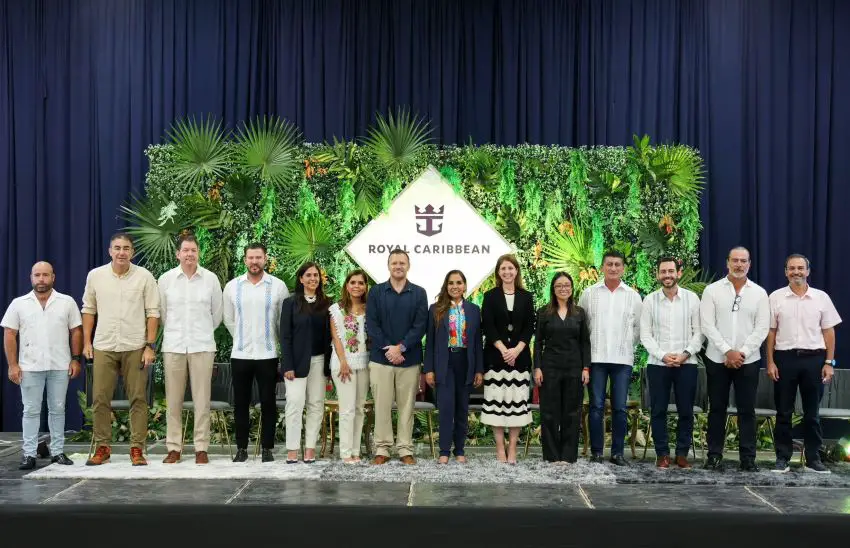 A line of people on a stage with the Royal Caribbean logo on a garden wall set up behind them.