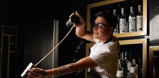 Bartender in a white shirt and black apron pours green liquid between the two cups of a metal drink shaker over an industrial bar sink. Behind him are bottles of various liquors on wooden shelving made to look like frames on a wall.