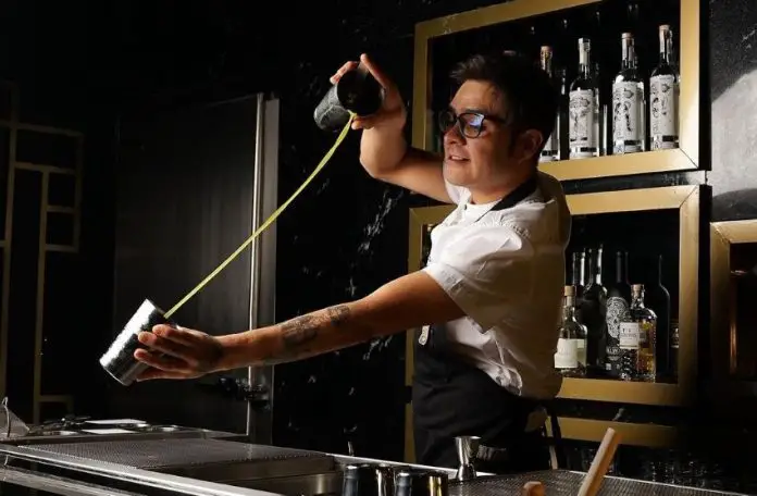 Bartender in a white shirt and black apron pours green liquid between the two cups of a metal drink shaker over an industrial bar sink. Behind him are bottles of various liquors on wooden shelving made to look like frames on a wall.