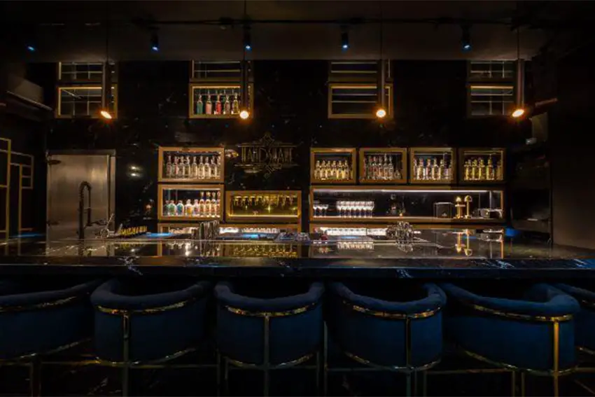 Dark bar counter decorated in brass and black with royal blue bar stools with lit shelves at the back wall filled with bottles of liquor.