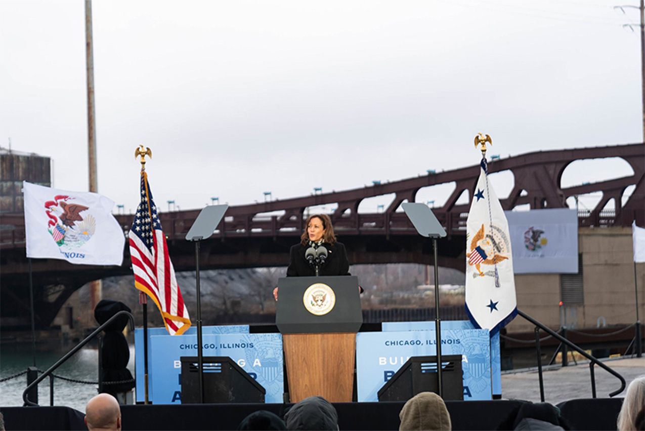 Kamala Harris at a podium at a rally in Chicago on an outdoor stage set up with a bridge in the background.