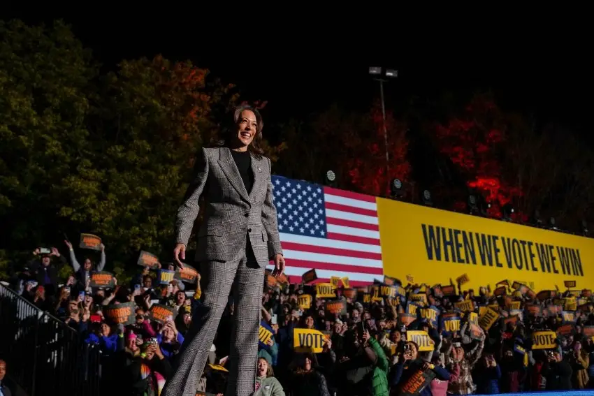 Kamala Harris standing on a stage with crowds of supporters behind and below her holding signs saying VOTE, with a yellow banner in the background with a US flag and the slogan When We Vote We Win.