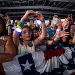 Kamala Harris supporters at a rally in the U.S. A older Latina woman and a young Latina girl are featured in this picture of a crowd behind a red, white and blue barrier. They are cheering and taking photos with their cell phones