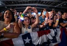 Kamala Harris supporters at a rally in the U.S. A older Latina woman and a young Latina girl are featured in this picture of a crowd behind a red, white and blue barrier. They are cheering and taking photos with their cell phones