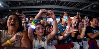 Kamala Harris supporters at a rally in the U.S. A older Latina woman and a young Latina girl are featured in this picture of a crowd behind a red, white and blue barrier. They are cheering and taking photos with their cell phones