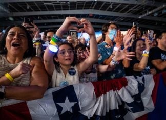 Kamala Harris supporters at a rally in the U.S. A older Latina woman and a young Latina girl are featured in this picture of a crowd behind a red, white and blue barrier. They are cheering and taking photos with their cell phones