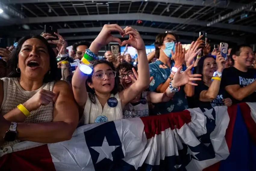 Kamala Harris supporters at a rally in the U.S. A older Latina woman and a young Latina girl are featured in this picture of a crowd behind a red, white and blue barrier. They are cheering and taking photos with their cell phones