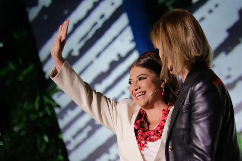 Clara Brugada wearing a headset and standing on a stage waving at the audience. Next to her is a woman in a leather jacket whose face cannot be seen.