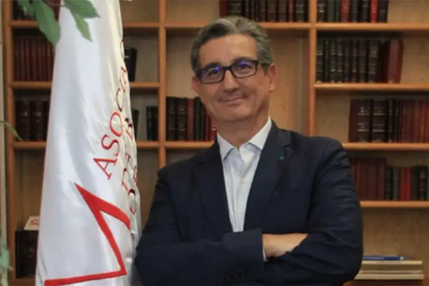 An elderly man in a dark suit, white shirt and thick black glasses standing with his arms folded in front of him next to the ABM flag.