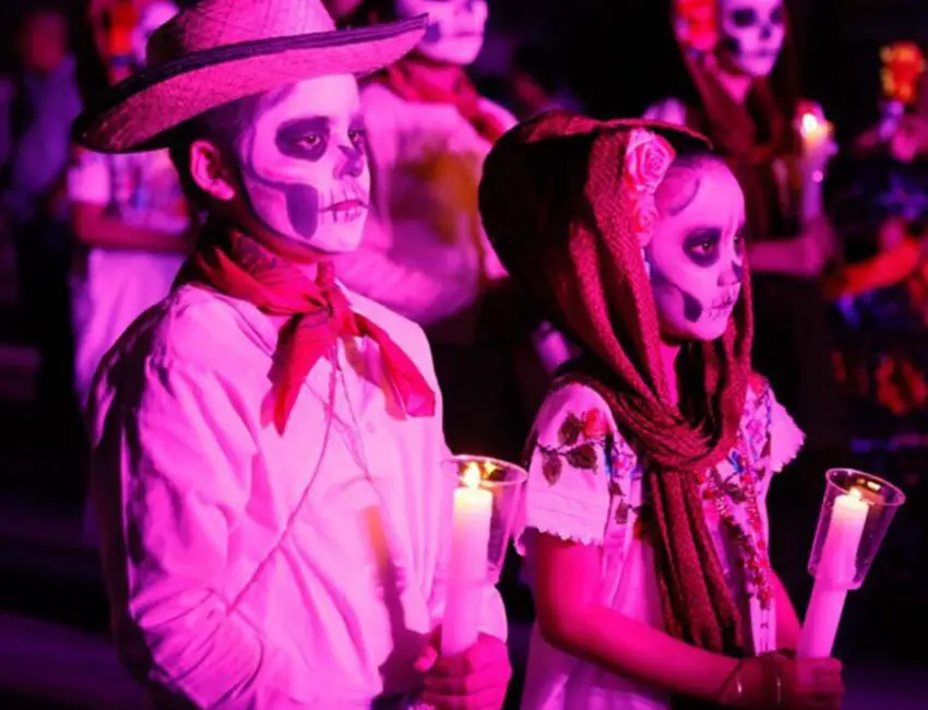 Un niño y una niña mexicanos maquillados al estilo del Día de los Muertos con ropa tradicional de Yucatán, sosteniendo gruesas velas blancas en sus manos como parte de una ceremonia nocturna del Día de los Muertos.