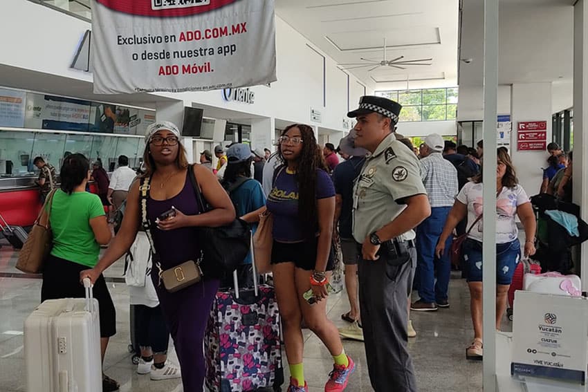 Travelers with suitcases walking through a Yucatan bus station with a National Guardsman watching them