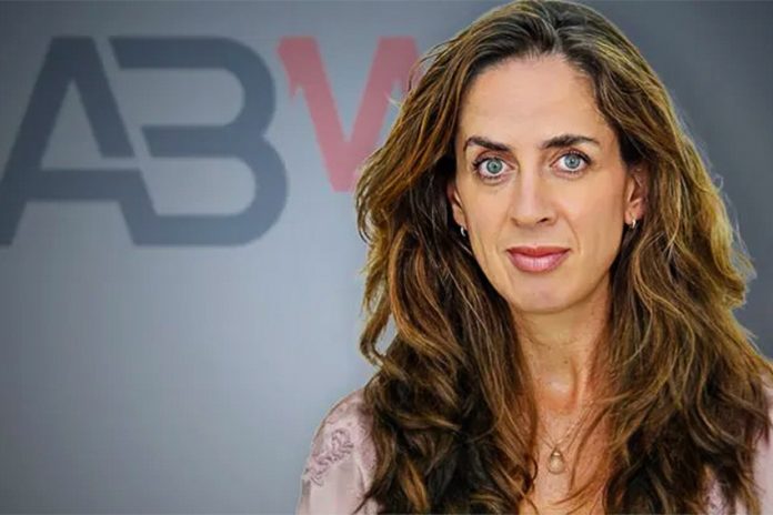 Woman with long wavy light brown hair staring into the camera, posing in front of a wall that has the letters ABM on it.