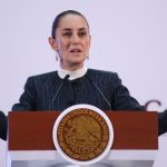 Claudia Sheinbaum at her press conference podium with her hands up and palms facing the floor in a gesture telling people to calm down.