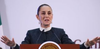 Claudia Sheinbaum at her press conference podium with her hands up and palms facing the floor in a gesture telling people to calm down.