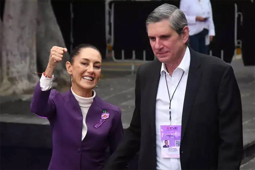 President Claudia Sheinbaum posing for a photo an an event with her husband, Jesús María Tarriba. Sheinbaum is smiling at the camera and has her right fist in the air while Tarriba looks off into the distance beyond the foreground.