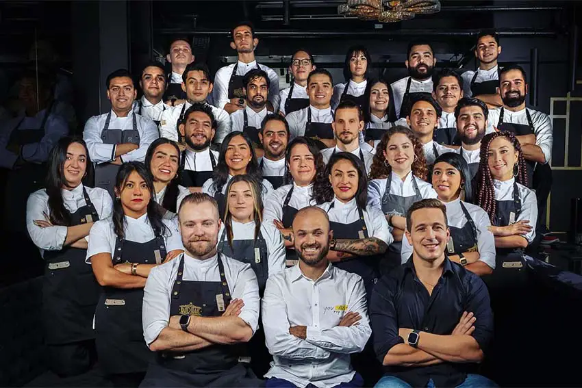 Bar workers posing for a photo in a pyramid setup with everyone wearing matching white shirts and black aprons except the two owners in front.