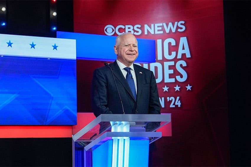 Gov. Tim Walz standing at a glass podium at the Vice Presidential Debate smiling in front of a projection wall for CBS News saying "America Decides"