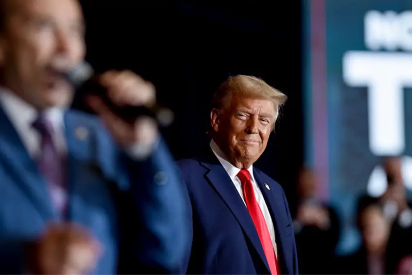 Donald Trump onstage at one of his rallies, looking on with a closemouthed smile as a male guest in a suit speaks in a microphone nearby.