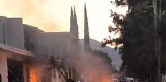 Smoke and flaming debris in the middle of a street where doors to buildings have been blown off. A silhouetted man stands in the street with his back to the camera.