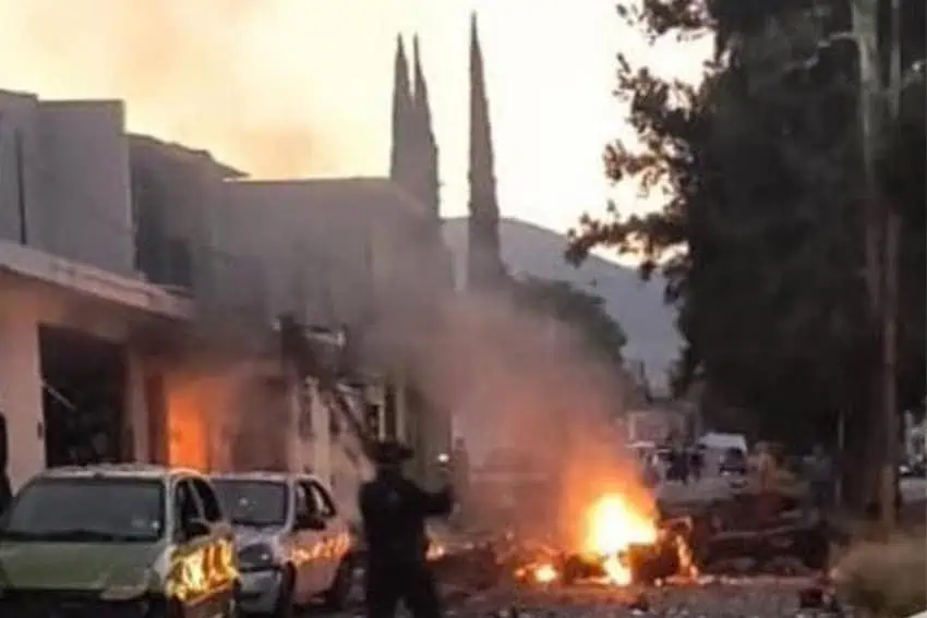 Smoke and flaming debris in the middle of a street where doors to buildings have been blown off. A silhouetted man stands in the street with his back to the camera.