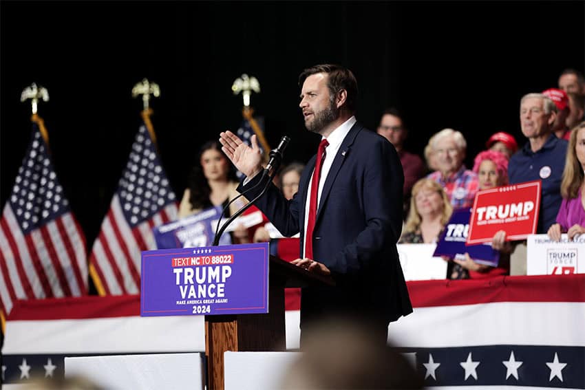 JD Vance standing at a podium at a Trump Vance rally with his hand in the air gesturing.