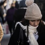 Young woman on a crowded street wearing a white, knitted winter hat with a pom pom and a white scarf and looking downward.