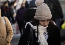 Young woman on a crowded street wearing a white, knitted winter hat with a pom pom and a white scarf and looking downward.