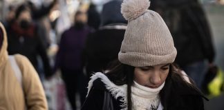 Young woman on a crowded street wearing a white, knitted winter hat with a pom pom and a white scarf and looking downward.