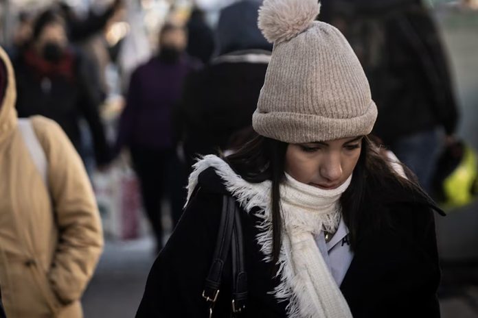 Young woman on a crowded street wearing a white, knitted winter hat with a pom pom and a white scarf and looking downward.