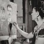 Black and white photo of Frida Kahlo looking at a painting she's working on of a portrait of her father, Guillermo Kahlo.