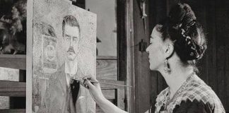 Black and white photo of Frida Kahlo looking at a painting she's working on of a portrait of her father, Guillermo Kahlo.