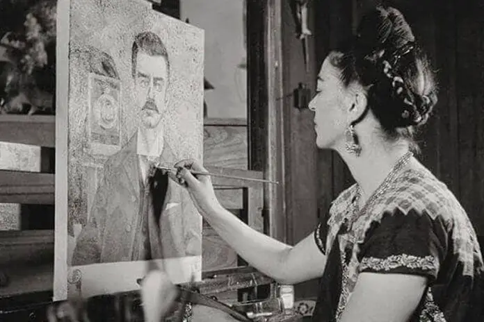 Black and white photo of Frida Kahlo looking at a painting she's working on of a portrait of her father, Guillermo Kahlo.