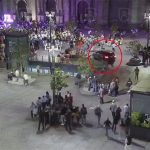 A car drives through a busy plaza in front of a cathedral, heading toward a crowd of people.