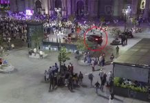 A car drives through a busy plaza in front of a cathedral, heading toward a crowd of people.